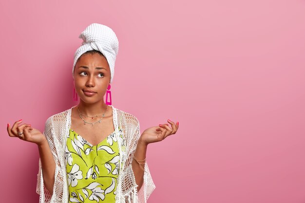 Photo of hesitant ethnic woman spreads hands sideways, feels hesitant, cannot decide what to wear for date, wears wrapped towel on head, faces dilemma, poses against pink wall with blank empty space