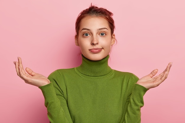 Photo of hesitant young European woman spreads palms, has confused expression, cannot make decision, wears green turtleneck, has unaware look, poses against pink background.