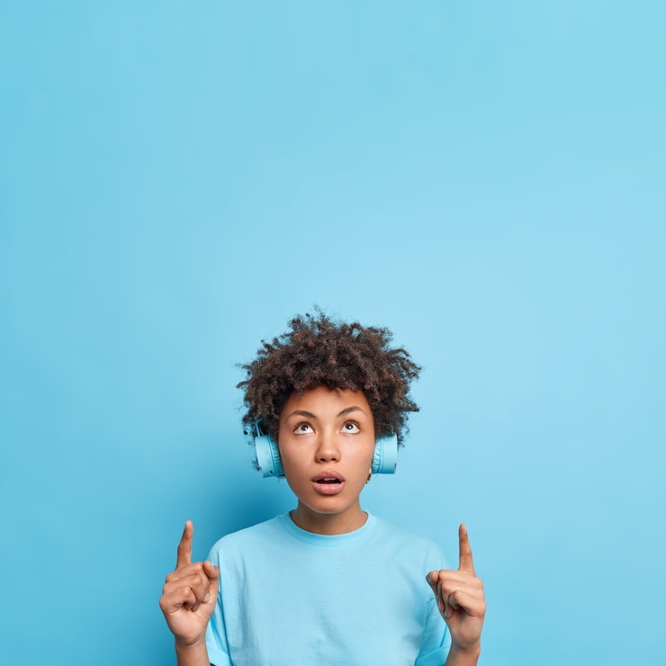 Free Photo photo of impressed curly haired woman says wow being wondered by something incredible points above on copy space shows advertisement up wears headphones on ears isolated on blue  wall.
