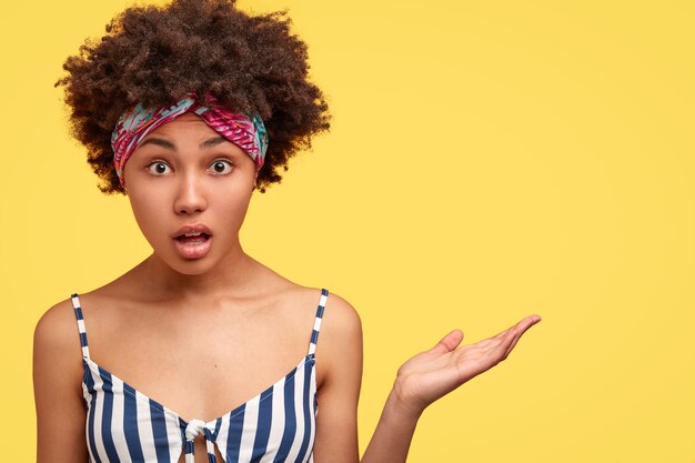 분개 놀란 젊은 아프리카 계 미국인 여자의 사진은 의아해 보이고, 노란색 공간에 대해 뭔가를 보여주고, 손을 들고, 줄무늬 탑을 입는다.