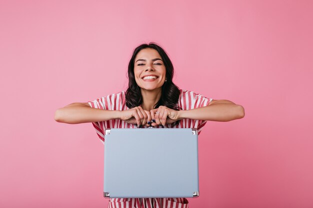 Photo indoors of positive dark-haired girl showing cute suitcase for money. Lady in great mood with snow-white smile looking.