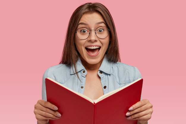 Free photo photo of intelligent european female wonk holds opened book, feels happy to read romantic story to end, feels excited with unexcpected event, wears jean jacket and round eyeglasses, stands indoor