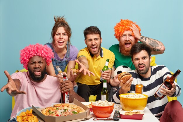 Photo of irritated football fans dissatisfied with result of game, look angrily at camera, drink beer, eat pizza, burger, popcorn