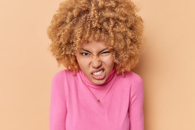 Free photo photo of irritated young curly haired woman clenches teeth with hateful expression smirks face dressed in casual pink turtleneck isolated over beige background. negative human emotions concept