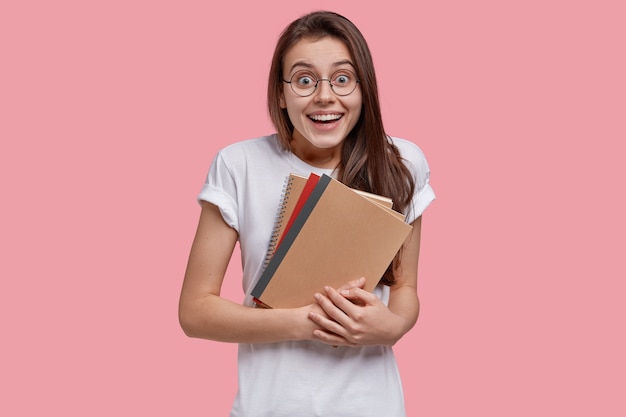 Photo of joyful dark haired woman carries spiral notepad, smiles broadly