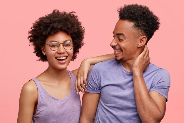 喜びに満ちた浅黒い肌の女性と男性の仲間の写真は、一緒に喜びを持ち、カジュアルな服を着て、前向きに笑い、ピンクの壁に立ち向かいます。幸せなアフリカ系アメリカ人の女性は男の肩に寄りかかる