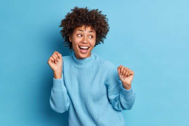 Photo of joyful dark skinned woman dances carefree keeps fists raised looks positively aside dressed in casual jumper moves 