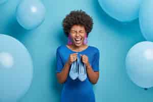 Free photo photo of joyful woman rejoices buying new outfit, holds blue stylish shoes to fit dress, dresses on special occasion, going to celebrate birthday. blue color prevails. fashion and clothing concept