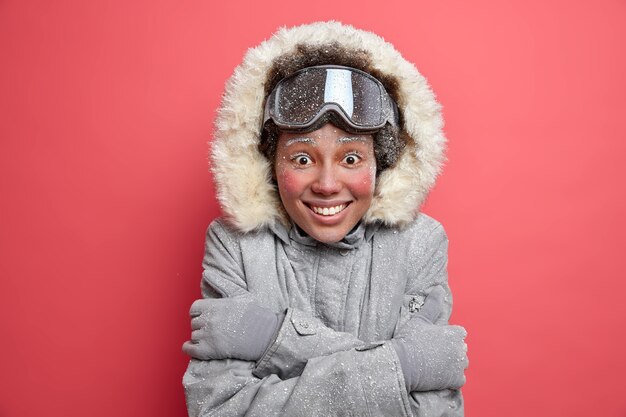 Photo of joyful woman with red cheeks covered by hoarfrost embraces herself wears warm thermo jacket.