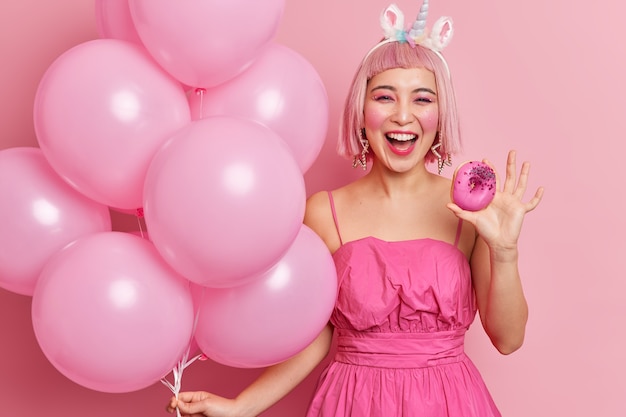 Photo of joyful young Asian woman has pink hair wears festive dress holds delicious glazed donut and bunch of inflated balloons enjoys party time 