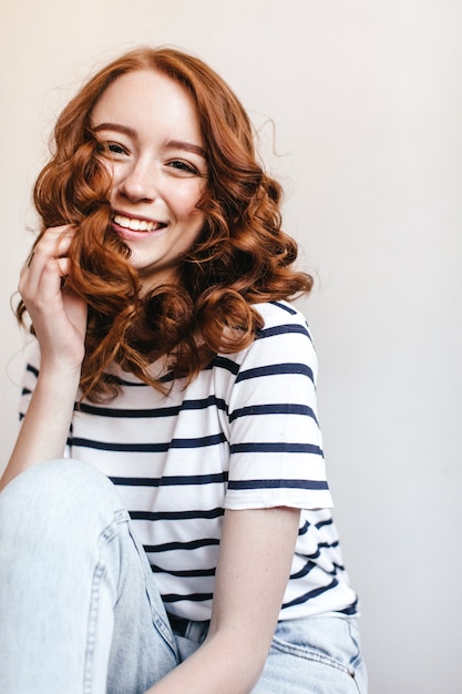 Photo of laughing jocund girl wears vintage blue jeans. Adorable ginger woman in striped t-shirt sitting near white wall.
