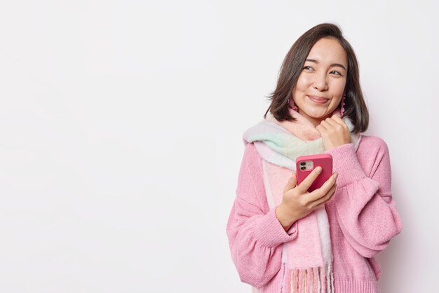 Photo of lovely brunette woman in warm sweater and scarf around neck uses mobile phone for surfing social networks waits for call from boyfriend looks dreamy aside isolated over white background