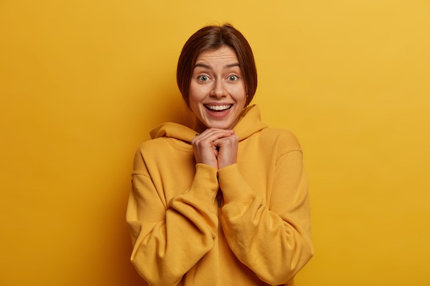 photo of lovely cheerful woman gazes happily, keeps hands under chin, spends free time in hilarious company, smiles satisfied and carefree, wears hoodie, poses against yellow  wall