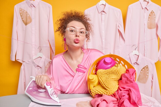 Photo of lovely curly haired busy housewife keeps lips folded poses near ironing board with laundry basket and steam electric iron wears transparent glasses and dressing gown has romantic mood