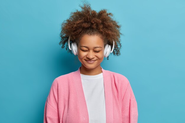 Photo of lovely dark skinned woman has upbeat mood wears headphones and listens pleasant music stands with closed eyes dressed in casual wear.