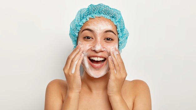 Photo of lovely female model with happy expression, washes face with foaming cleanser, wears wateproof showercap, pampers skin, stands shirtless, looks straightly. Facial treatment