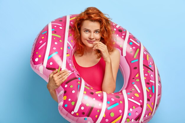 Photo of lovely ginger woman looks happily, poses inside of rubber swim ring, dressed in swimwear, rests at seaside