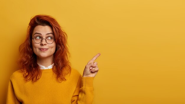 Photo of lovely ginger woman points index finger aside, demonstrates promo on right, looks with interesting expression, has wavy red hair