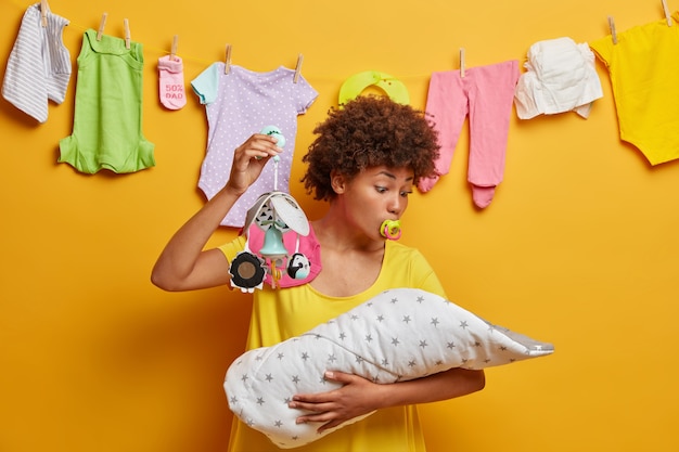 Photo of lovely mommy looks at baby and tries to sooth naughtly newborn, shows mobile and sucks nipple, nurses infant, plays with little daughter, stands over yellow wall with washed clothes