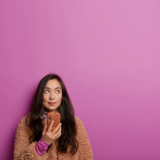 La foto di una bella donna premurosa sta con il cellulare moderno, guarda da qualche parte, ha un'idea interessante, riceve una proposta curiosa, vestita con un cappotto marrone