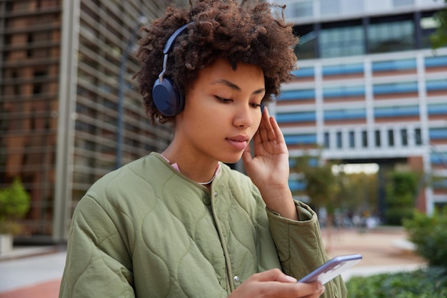 Photo of lovely woman with curly bushy hair listens media player during free time holds mobile phone checks song from playlist