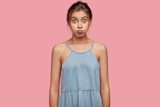 Photo of lovely young girl never looses sense of humour, blows cheeks, has fun and makes grimace