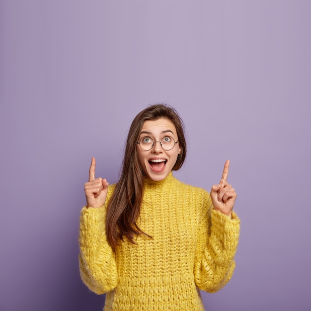 Photo of lovely young woman looks gladfully, points above on free space, wears casual yellow sweater, round spectacles