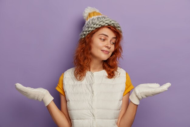 ミレニアル世代の女の子の写真は、手のひらを横に立って、何かを持っているふりをして、2つのアイテムから選択し、帽子、白い手袋を着用し、冬休みを楽しんで、紫色の背景で隔離
