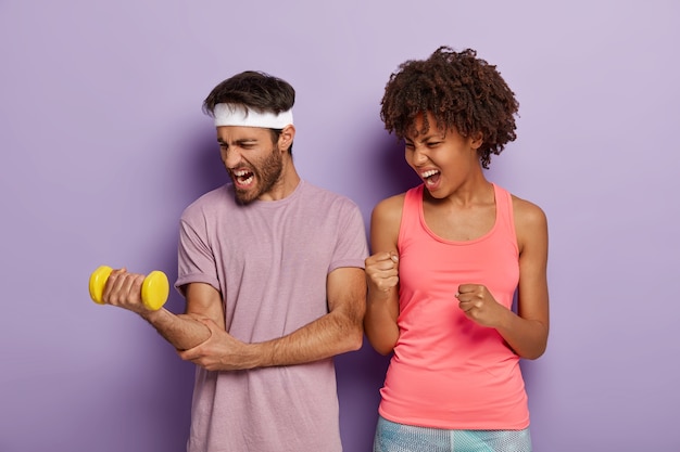 Foto gratuita la foto di un uomo caucasico motivato alza il braccio con un manubrio pesante, lavora sui muscoli, una donna dalla pelle scura e solidale stringe i pugni e grida, crede nel successo del fidanzato. risultati sportivi