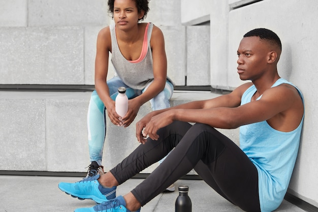 Photo of motivated dark skinned man and his female companion, pose together at stairs, have break after outdoor cardio training, drink water, have athletic body shape, run up. Lifestyle concept