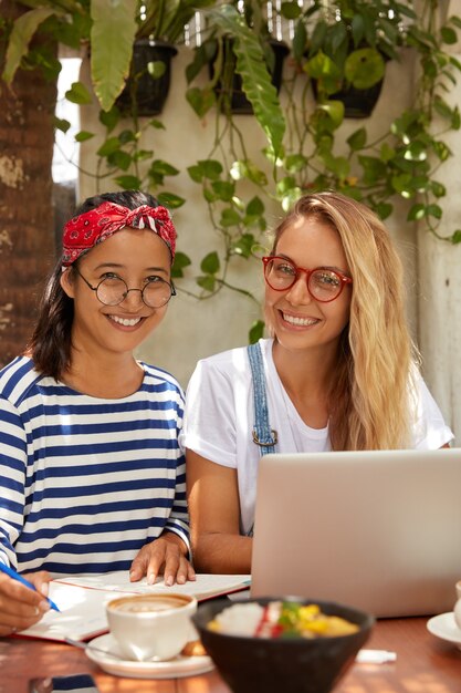 Photo of multiethnic women make research work together: blonde female in spectacles searches information in internet while her companion writes in notebook