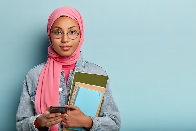 Photo of Muslim student carries notepad for notes, holds modern cellular, creats new publication in social networks, covers head with veil according to religious rules, chats with groupmates online