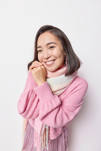 Photo of nice looking Asian woman smiles gently keeps hands near face has dreamy expression thinks about something pleasant wears pink jumper and scarf around neck isolated over white background