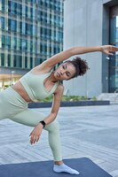 photo of active sporty curly haired woman leans aside keeps arm raised up dressed in activewear poses on fitness mat against urban background being in good physical shape does bend exercise