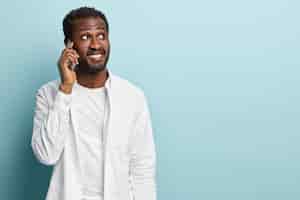 無料写真 黒人の流行に敏感な男の写真は、電話で会話し、携帯電話を耳の近くに持ち、友人にニュースを伝え、集中し、白いシャツを着ています。成功した実業家は携帯電話を介して予定を立てます