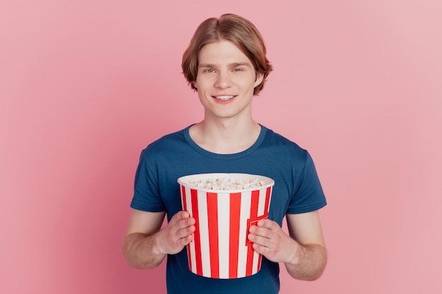 쾌활한 사람이 팝콘 종이 상자를 들고 있는 사진은 행복한 긍정적인 미소로 영화관에서 격리된 분홍색 배경을 보고 있습니다.