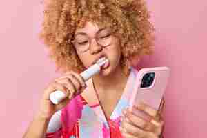 무료 사진 곱슬머리 여성이 치약을 사용하여 이를 닦는 사진은 분홍색 배경에 격리된 스마트폰을 통해 자신의 사진을 만듭니다. 치과 치료 및 일상적인 위생 개념