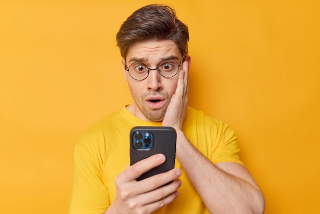 Фотография красивого темноволосого взрослого мужчины с удивленным выражением лица смотрит на экран смартфона, держит руку на щеке, реагирует на шокирующие новости, одетый в повседневную футболку, изолированную на желтом фоне Premium Фотографии