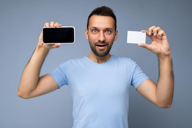 Фотография красивого молодого небритого брюнет с бородой в повседневной синей футболке, изолированной на синем фоне, держащей и использующей мобильный телефон и кредитную карту для оплаты, глядя в камеру