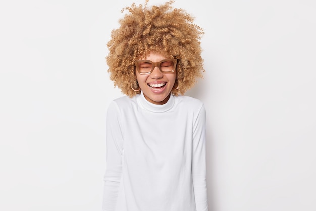 Бесплатное фото Фотография обрадованной кудрявой европейской женщины смеется, улыбается зубами, держит глаза закрытыми, носит очки и водолазку, изолированные на белом фоне. концепция счастливых эмоций и чувств