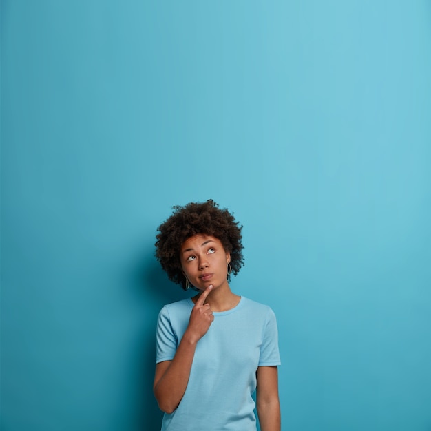 Бесплатное фото Фотография задумчивой темнокожей женщины, которая держит палец на подбородке, сосредоточена наверху и о чем-то размышляет, носит футболку, позирует у синей стены, пустое место для вашей рекламы или информации