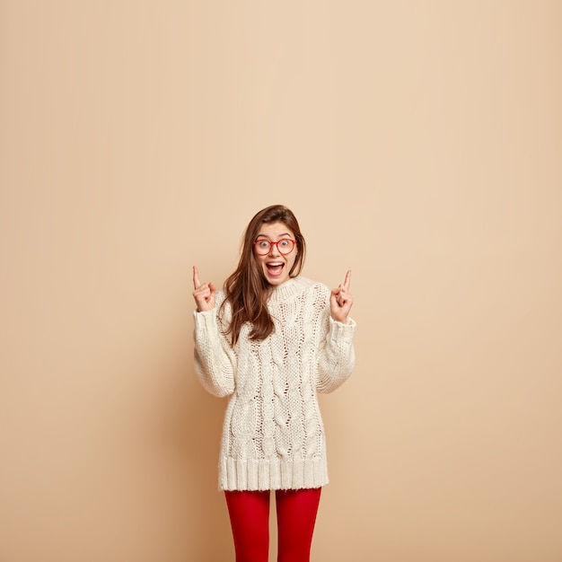 Бесплатное фото Фотография позитивно обрадованной самки с широко открытым ртом, кричит от возбуждения, указывает вверх на свободное пространство, одетая в белый джемпер, прозрачные очки, изолирована на бежевой стене