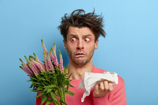 Бесплатное фото Фотография ошеломленного человека смотрит на растение широко открытыми глазами, у него растрепанные волосы, он держит салфетку, постоянно чихает, аллергическая реакция на пыльцу и цветение, чувствует себя больным и измученным из-за болезни