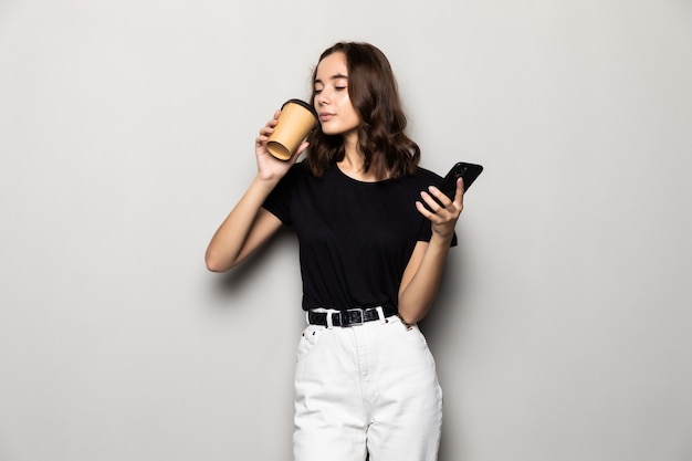 Фотография успешной женщины в формальной одежде, стоящей со смартфоном и кофе на вынос в руках, изолированной над серым