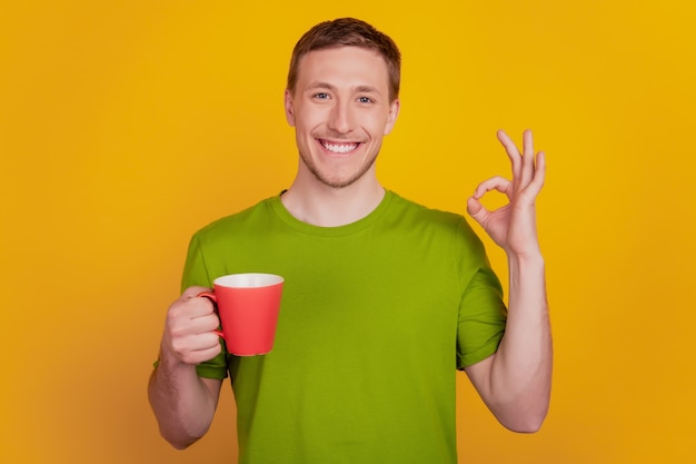 젊은 남자의 행복한 긍정적인 미소 음료 커피 컵의 사진은 노란색 배경 위에 격리된 괜찮은 표시를 보여줍니다.