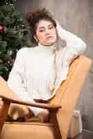 무료 사진 크리스마스 트리 근처의 편안한 의자에 앉아 있는 젊은 여성의 사진