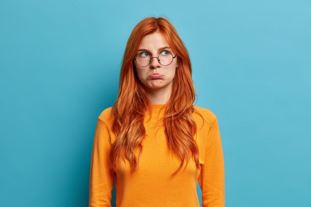Photo of offended redhead young European woman has moody disappointed look purses lips and looks away discontent by hearing unpleasant words wears round spectacles casual jumper.