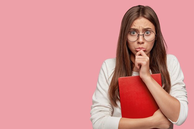 Photo of offended student purses lower lip with negative emotions, being dissatisfied with bad mark, holds red textbook