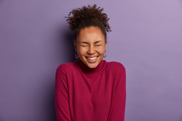 Photo of optimistic curly woman with dark skin, closes eyes and smiles broadly, shows white teeth, feels overjoyed, expresses good emotions, hears hilarious joke, isolated over purple  wall