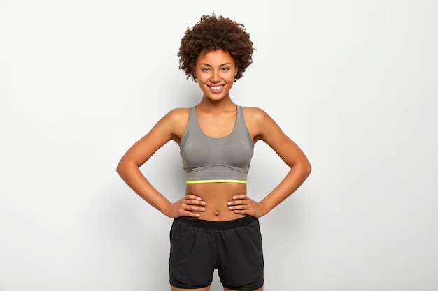 Free photo photo of optimistic dark skinned sporty woman keeps hands on waist, smiles happily, dressed in sport bra and black shorts, isolated over white background